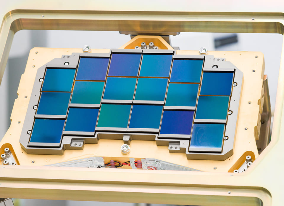 Eighteen square infrared detectors in a stepped mosaic, each one iridescent blue and green, seated in a gold engineering test frame.