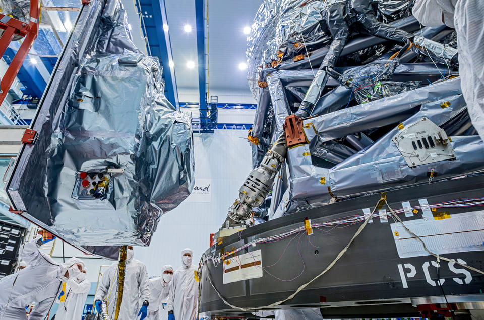 The Wide Field Instrument, wrapped in silver thermal blanketing and suspended from a lifting fixture, held beside the strut work of the Roman payload structure, with technicians in clean room suits standing below it.